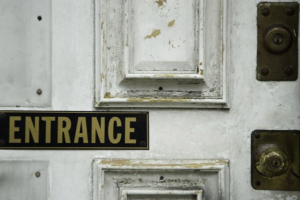 An old, worn white door has peeling paint and a metal
'Entrance' sign.