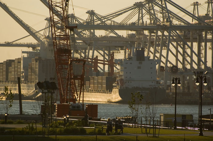 A couple sits on a bench, with a container ship and several
cranes in the background.