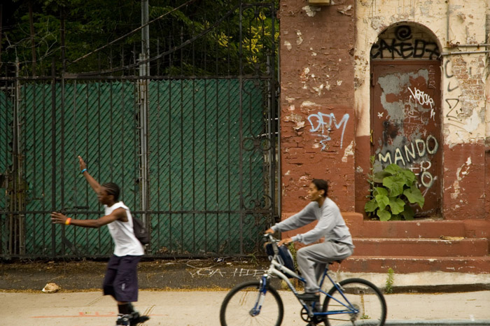 Two people go past a fence and graffiti-covered wall.