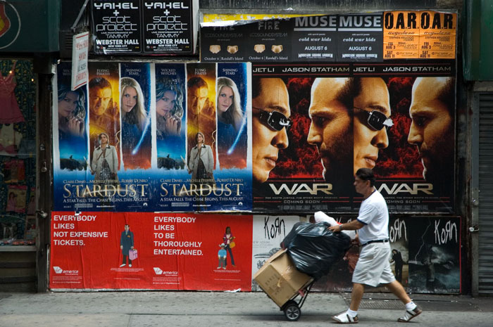 A man wheels a loaded hand truck past repetitive ad
posters.
