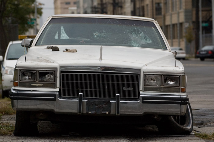 A Cadillac has a smashed windshield and other signs of ruin.