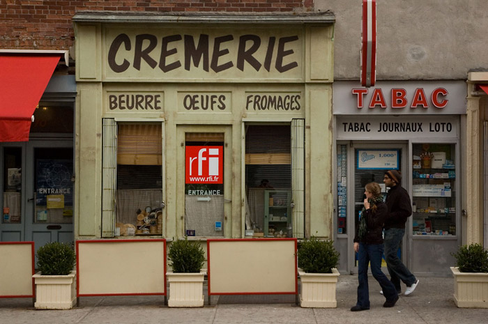 Two people walk past 'Cremerie' and 'Tabac' storefronts.