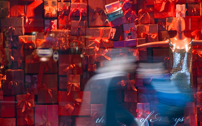 Two people walk quickly past a store window filled with wrapped red presents and a mannequin.