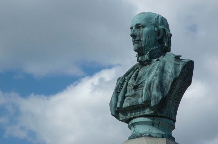 A bust of Horace Greeley, copper green and corroded, stands out against a brilliant blue clouded sky.