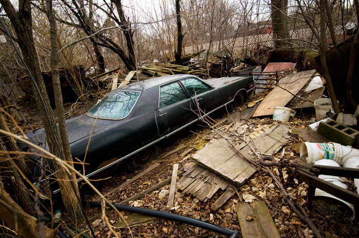 An old car sits locked away in a yard with a variety of junk and broken timber.
