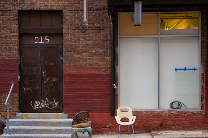 A cream-colored, plasticchair sits out in front of a red brick building with a black door.