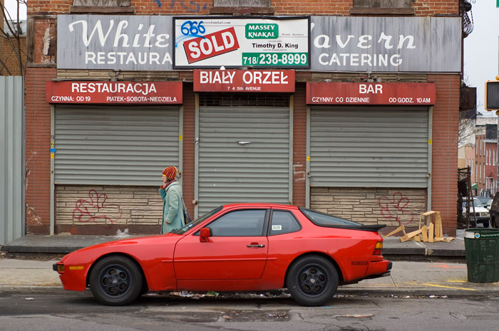 A woman in a red knit cap walks past a restaurant with red canopies and a red sign, as well as a red sports car. Oh, and red graffiti, too.