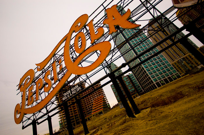 A huge, red neon sign, advertising 'Pepsi Cola' stands in front of high-rise apartment buildings.