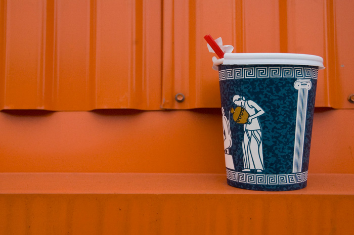 A paper coffee cup with Greek designs shows an attendant pouring oils or something over her mistress. The blue and white cup is against an orange wall.