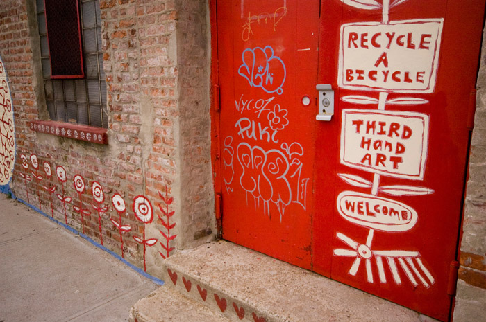A metal double door has been playfully painted in red, offering bicycle recycling, art, and a welcome. Red flowers have been painted on the brick wall.