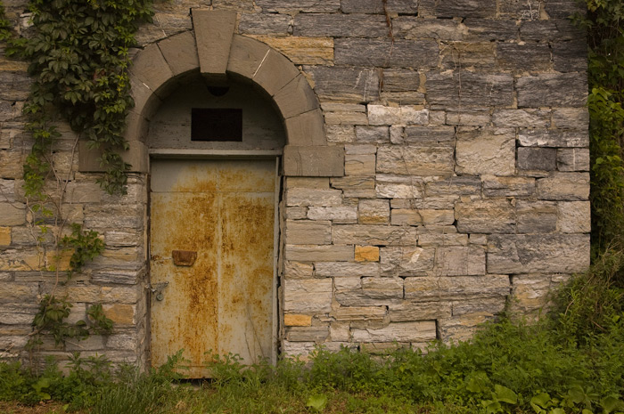 A building is made of rough-hewn stone. It has a rusted door, with a stone arch over its top.