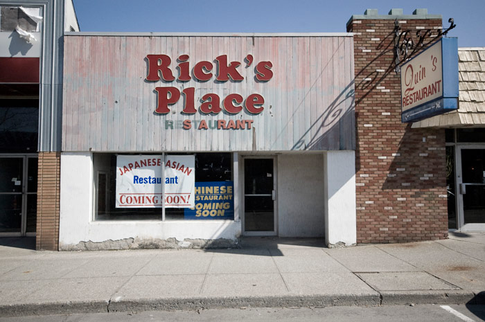 An empty storefront has signs left over from two former restaurants, and both signs are in bad shape. Signs in the window promise a new restaurant coming, but it promises too many cuisines