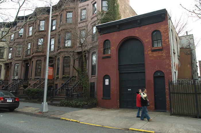 Two people walk past a short building which was a stable, indicated by its very tall center entrance.