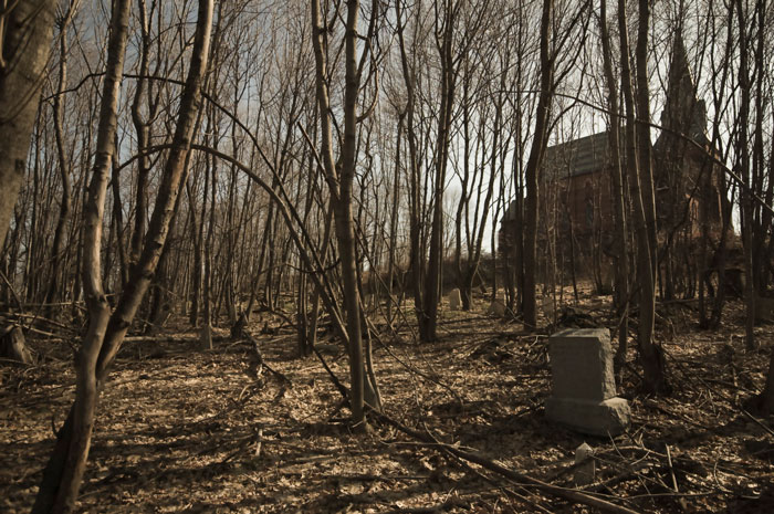 A handful of tombstones sit among bare trees in the winter; up the hill is a silhouette of a church.