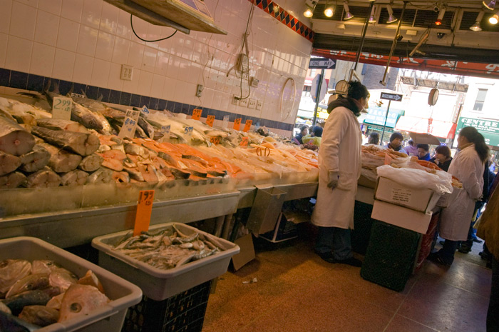 Looking out from within a Chinatown fish store, bins of fish in ice are on the left; the floor is tiles, and the attendees look out to the street.