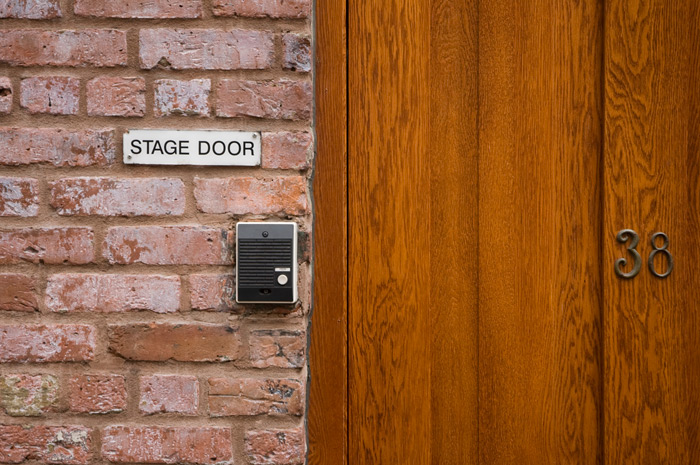 A sign on a brick wall reads 'Stage Door'. There is also an intercom buzzer, and a wooden door to the right