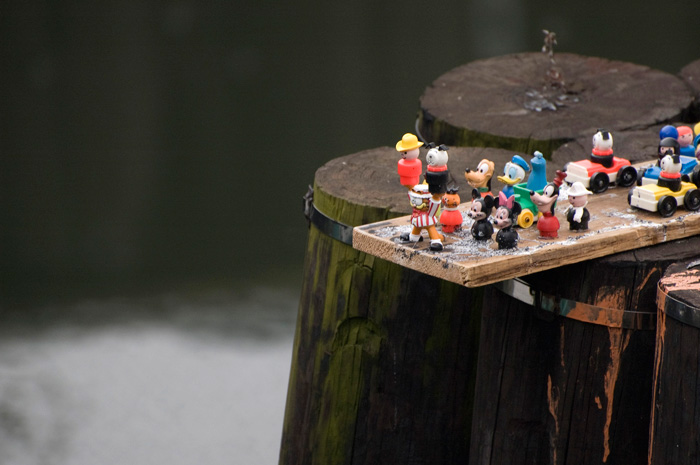 Over a dozen small toys (including Garfield the cat, Donald Duck, Mickey and Minnie Mouse, and Goofy) have been placed on a board, as if preparing to march into a canal.
