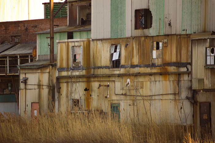 A building with discolored fiberglas siding has broken windows and is surrounded by overgrown weeds.