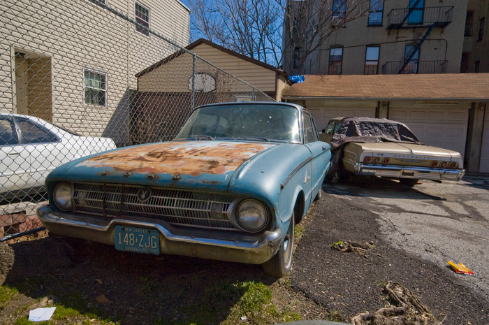 A rusty blue car sits in a front yard. Behind it is a second junked car, with a tap covering its missing back window.