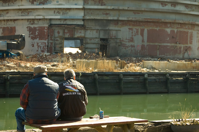 Two men, their backs to the camera, sit on a bench and watch the demoliton of a factory in the background.