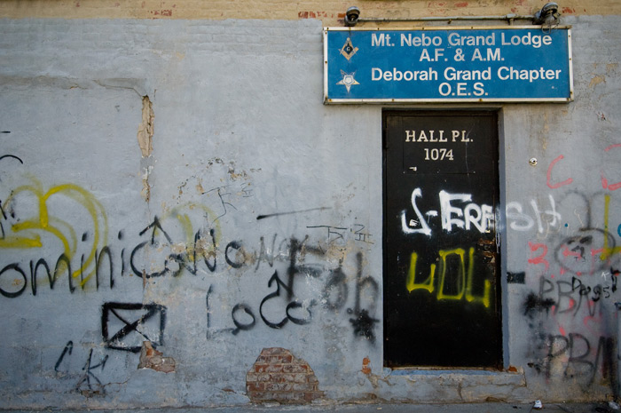 Spraypaint covers a wall of a building without a roof; in the wall is a black door, and above it a sign announcing its former role as a Masonic lodge.