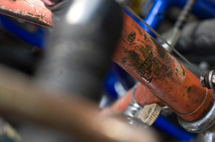 A tangle of bicycle frames, showing their age... An orange one sticks out, and its decal no longer shows ita brand very well.