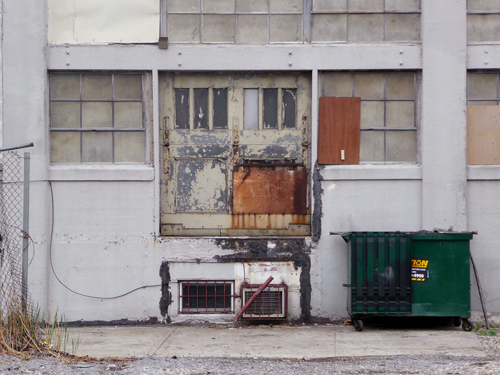 An industrial building has a double-door loading bay, with window panes in the doors. It's rusting. To its right is a green dumpster.