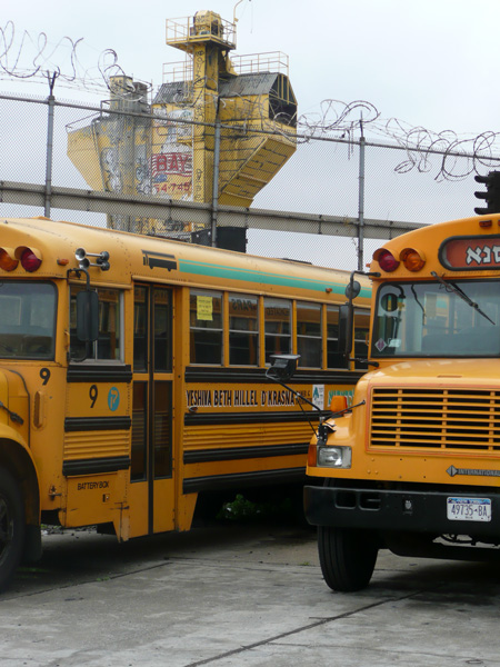 Yellow school buses sit in a lot; behind them rises the yellow tower of a concrete factory.