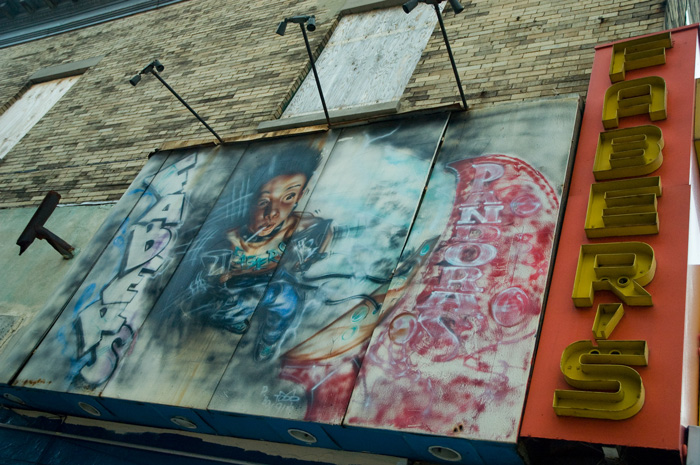 A faded mural shows a youth opening a Pandora's Box of drugs.  To its right is a neon sign that no longer works.