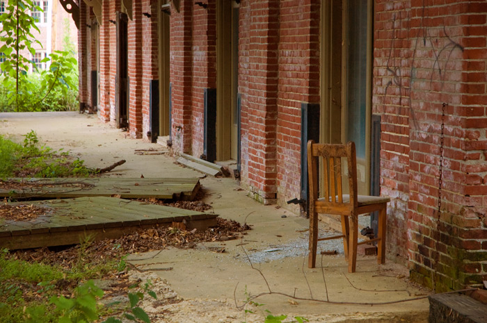 A concrete walkway next to a brick building complex is overgrown with weeds, and has a solitary chair by the wall.