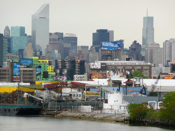The Newtown Creek and industrial buildings are in the foreground, with Manhattan skyscrapers in the background.