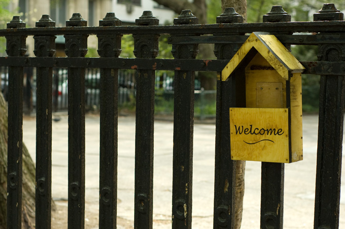 An iron fence outside a Quaker meeting hall has a yellow bulletin box hanging on it, painted with the word 'Welcome.'