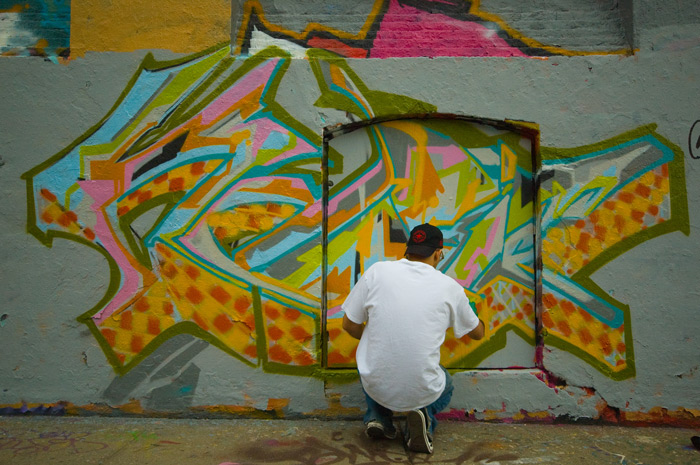 A man in jeans, a white T shirt, and a baseball cap squats while painting 'street art' on the side of a building covered with street art.
