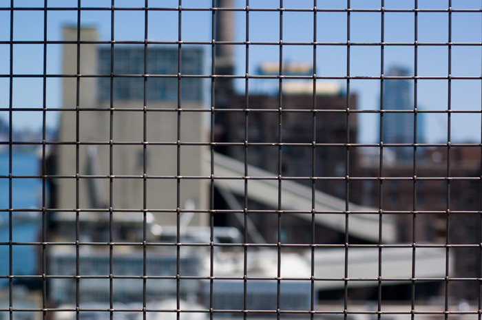 The outline of a factory can be seen through the square grid of a wire fence.