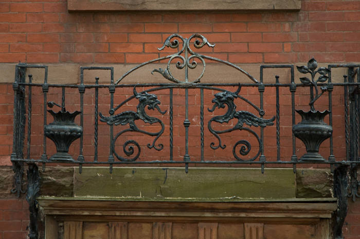 A brick building is ornamented with a cast-iron railing consisting of a pair of gryphons facing each other.