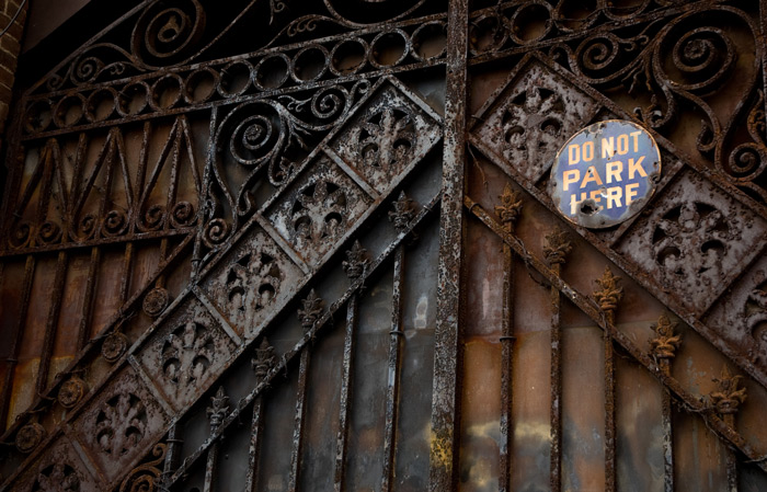 A driveway's two iron doors, with elaborate iron work, have several layers of paint, and a sign reading 'Do Not Park Here.'