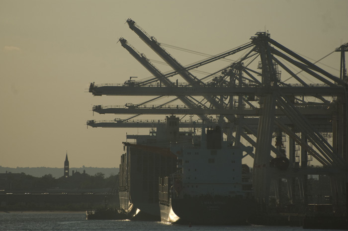 A container ship is docked under a series of shipping cranes, and a steeple stands tall in the distance.