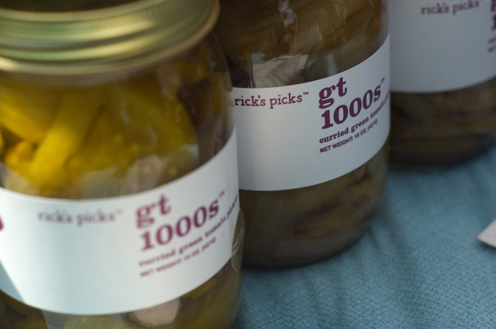 Jars of pickled green tomatos stand on a blue table cloth, for sale.