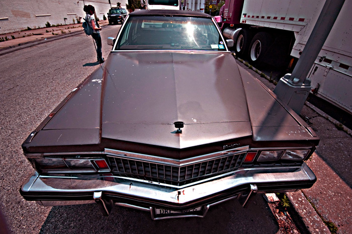 The photo shows a front end view of a Cadillac, going back to the windshield. Huge trucks are parked nearby.