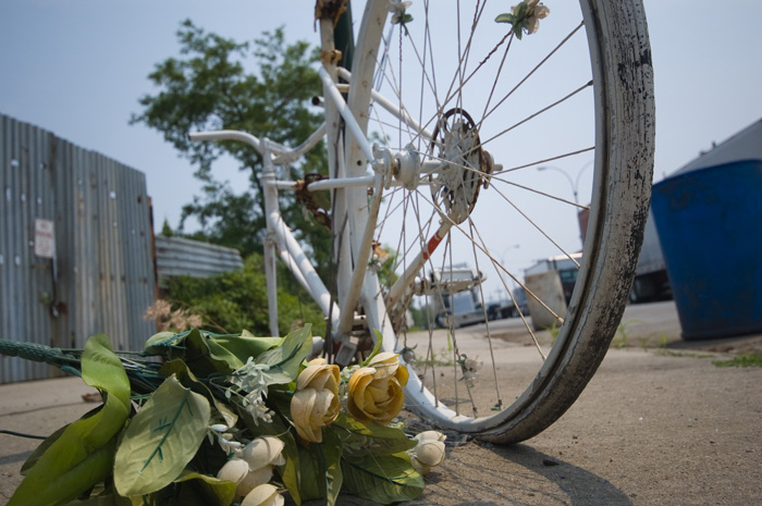 A faded bouquet of artificial flowers lays on the sidewalk, next to the rear wheel of a bicycle painted white and locked to a street sign.