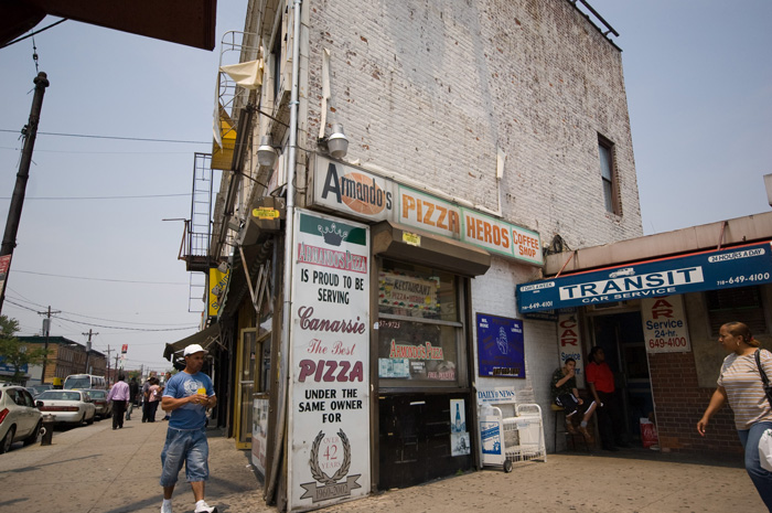 A corner pizza parlor stands by an active street and a subway exit.