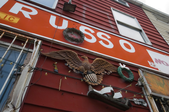 A red building with aluminum siding, now empty, has old decorations such as an American Eagle, a wreath, and signs announcing notary services.
