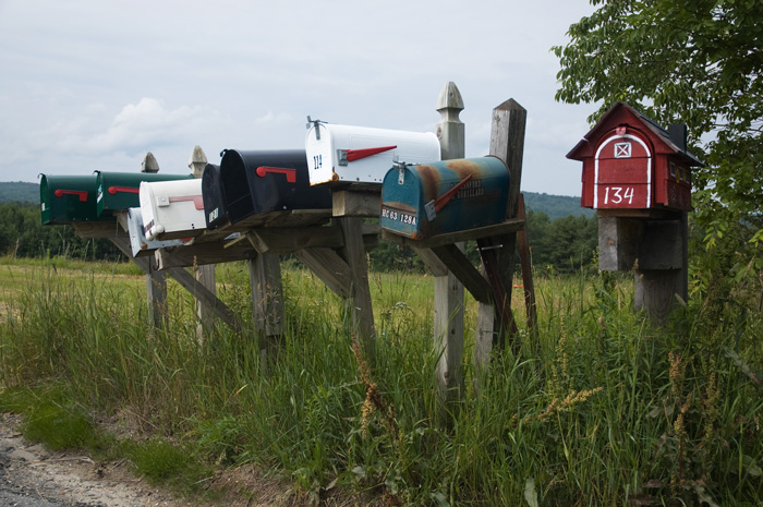 Nine mailboxes of different designs stand on wooden posts by the side of a road, next to an open field.