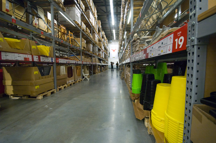 An aisle in an Ikea store's warehousing section is stacked with boxed items, and two people are at the far end.