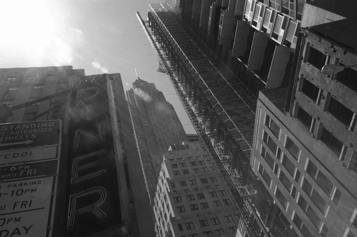 Sun streams down between tall skyscrapers, highlighting a diner's neon sign.