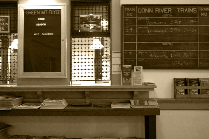 A ticket window in an old train station is next to racks of brochures and a chalk board showing schedules.