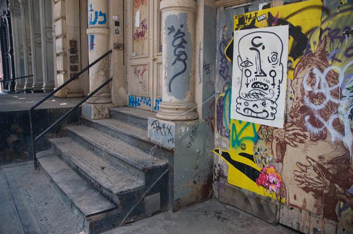 Beside the steps of a Soho building, pillars have been covered with graffiti, and a nearby wall has several prints pasted up.