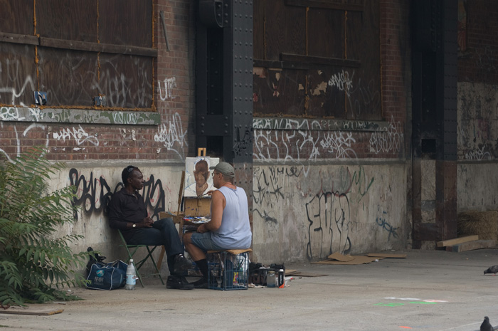 A portait is being painted; the subject is sitting with his back to a wall covered in graffiti, and the painter sits on top of a milk crate, in front of his canvas.