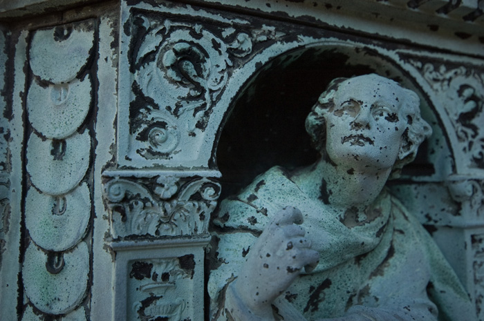 A face stares up from the ornate base of a metal pillar; all have corroded and are now bluish green.