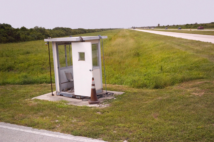 An empty security booth stands by the side of a road, and cars zoom to and fro.